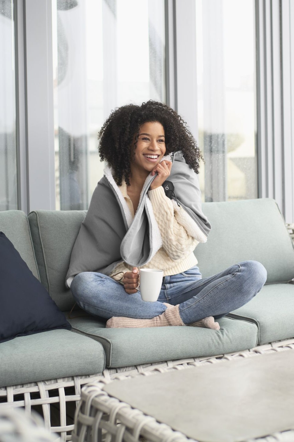 Eine Frau sitzt eingekuschelt in eine beheizte Wärmedecke auf einem Outdoor-Sofa und hält eine Tasse in der Hand.